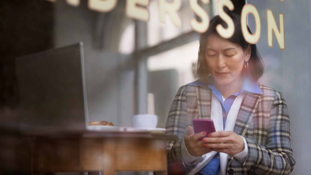 Person som sitter i en restaurang emd en telefon i handen och en dator framför sig på bordet.