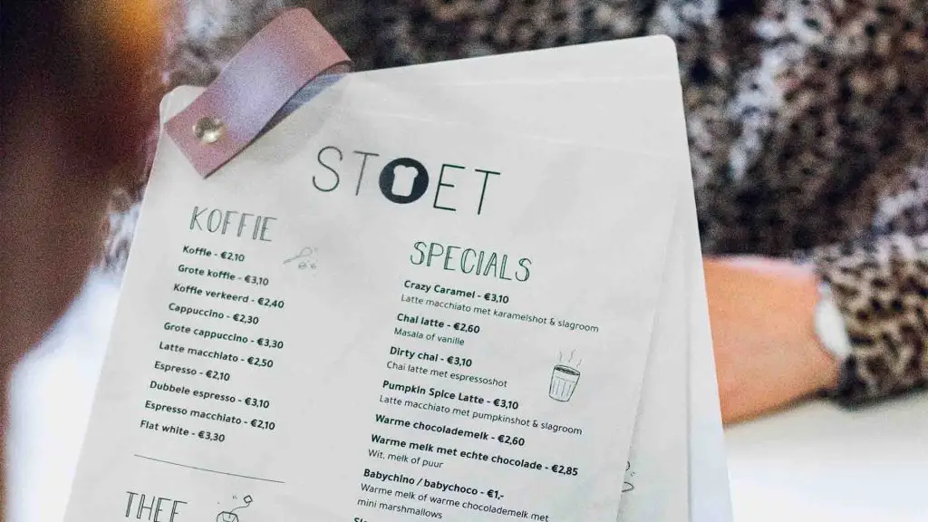 En restaurang meny som en person håller i och kollar på. Mitt emot personen som håller menyn sitter en annan person i prickig skjorta