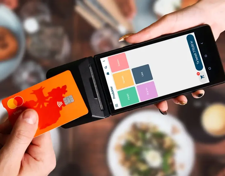 En person som betalar med oranget bankkort på en kortterminal med inbyggd kassasystem från Onslip. Händerna och kortterminalen som syns i bilden sker ovanför ett matbord som är oskarpt i bakgrunden. I bakgrunden ser man tallrikar med mat på.